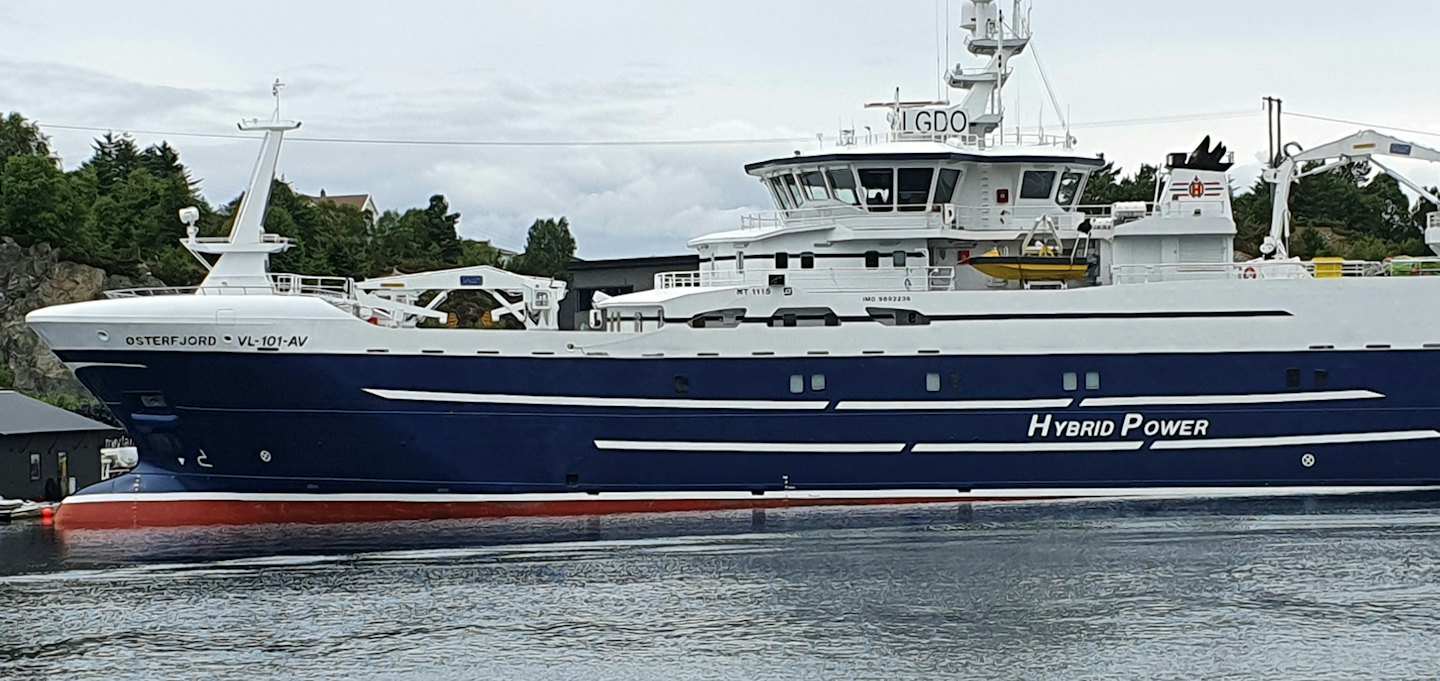 A hybrid power work vessel floats on calm water near land.